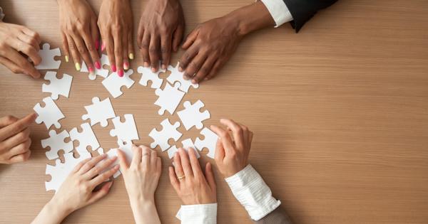 Five people's hands are on top of a wooden table, working together to put puzzle pieces together.
