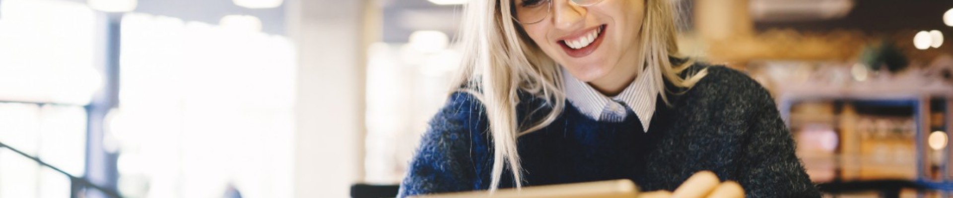 A young woman sits in a modern, well-lit library, smiling at something she's looking at on a tablet.