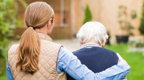 A mid-shot of two women walking away from us in a garden. One is wearing a cream gilet over a light blue shirt, and has her arm around the other woman, who is smaller and elderly. The older woman has white hair and wears a navy blue cardigan.