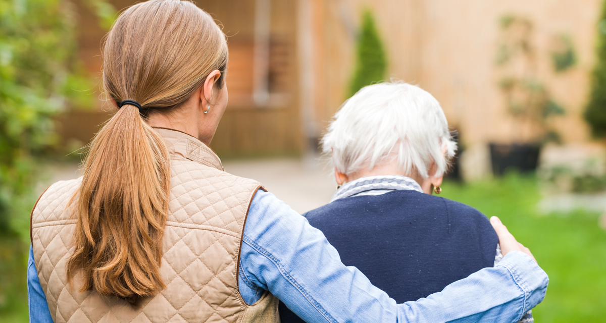 A mid-shot of two women walking away from us in a garden. One is wearing a cream gilet over a light blue shirt, and has her arm around the other woman, who is smaller and elderly. The older woman has white hair and wears a navy blue cardigan.