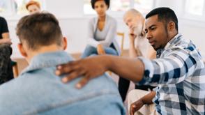 A group of people sit on chairs in a circle. The man closest to the camera has his head down, and the man next to him is patting him on the back to provide comfort.